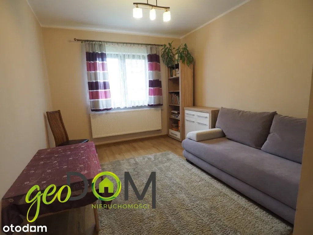 This living space is furnished with a comfortable grey sofa and a plush rug, creating a cozy atmosphere. A wooden dining table with a patterned tablecloth sits to the left, acco...