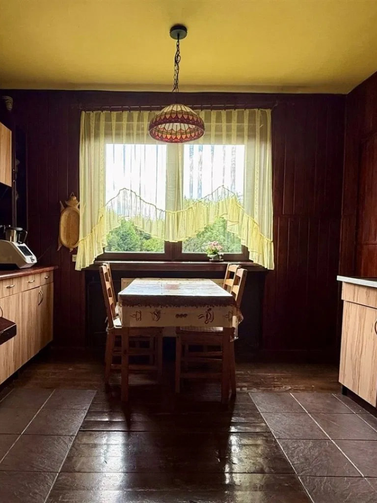 This rustic kitchen features dark wood paneling on the walls and a warm yellow ceiling, creating a cozy atmosphere. A simple wooden table with two chairs is set in front of a la...