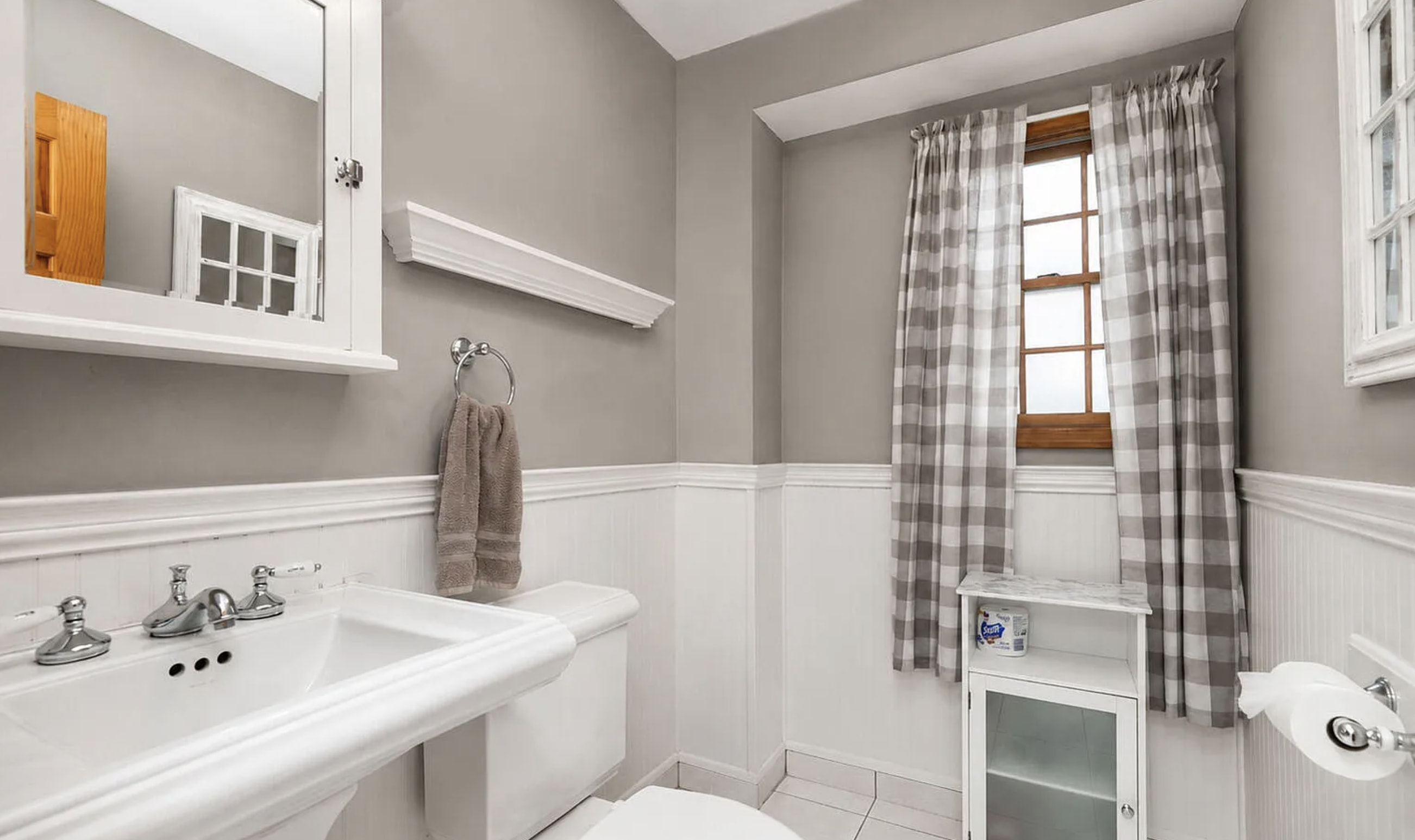 This bright and clean bathroom features a pedestal sink with chrome fixtures and a white porcelain toilet. The walls are painted a soothing neutral gray above white beadboard wa...