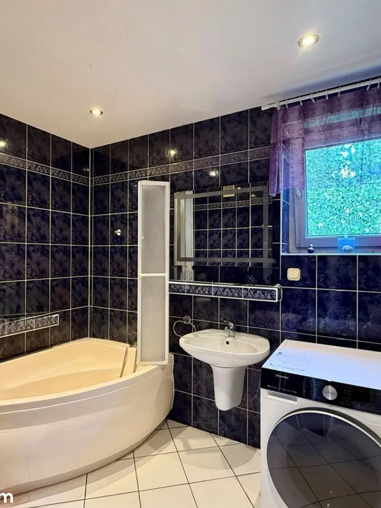 This bathroom features dark, glossy tilework on the walls, creating a dramatic and modern aesthetic. A spacious, corner bathtub with a curved design is the focal point, compleme...